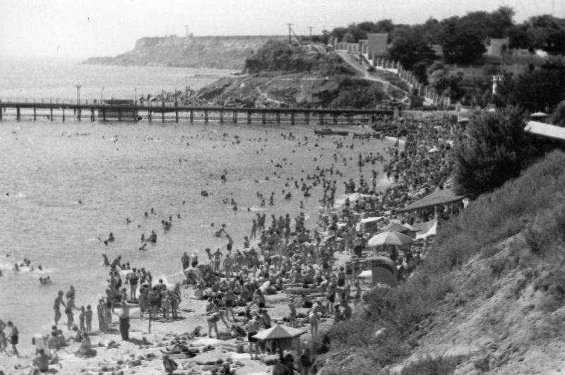 A beach panorama in high season: people on the sand and in the water, a wooden pier in the sea, a grassy slope on the right and a headland in the distance