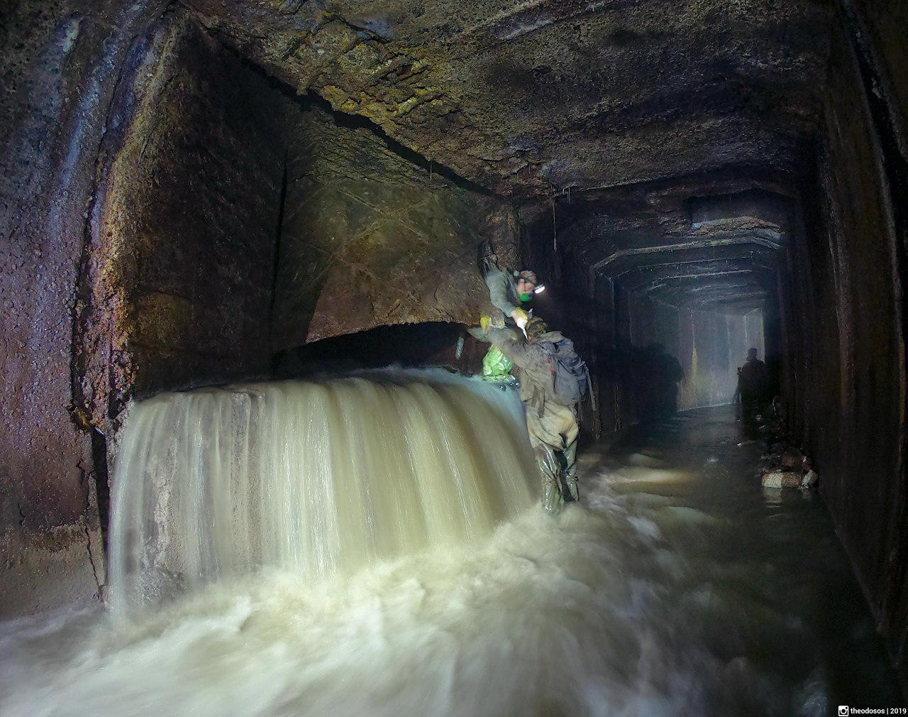 An underground tunnel with a stream of water: a waterfall over a sill, two people in head torches